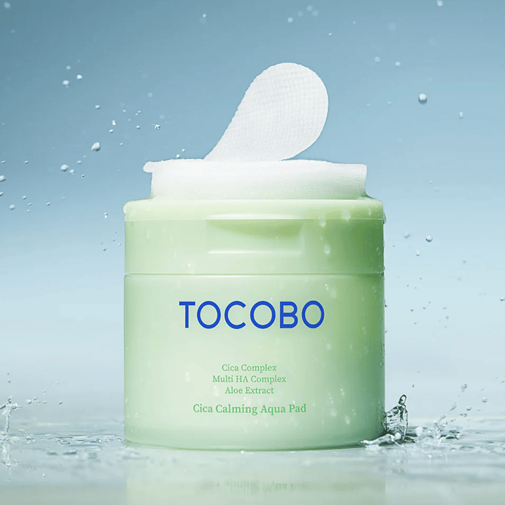 Åben krukke med TOCOBO Cica Calming Aqua Pad, hvor en toner pad stikker op af emballagen. Vanddråber rundt om produktet forstærker følelsen af intensiv fugt og friskhed.