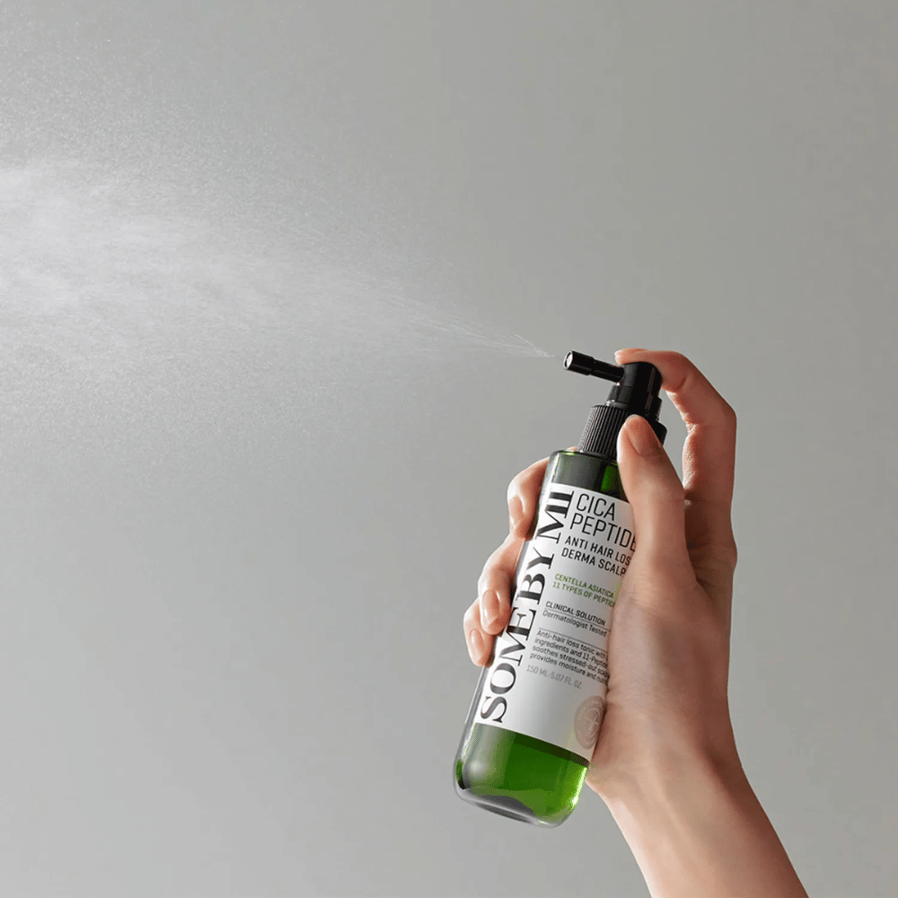 Hånd der sprayer SOME BY MI Cica Peptide Anti Hair Loss Derma Scalp Tonic fra en grøn flaske med sort mundstykke og hvid etiket mod grå baggrund.
