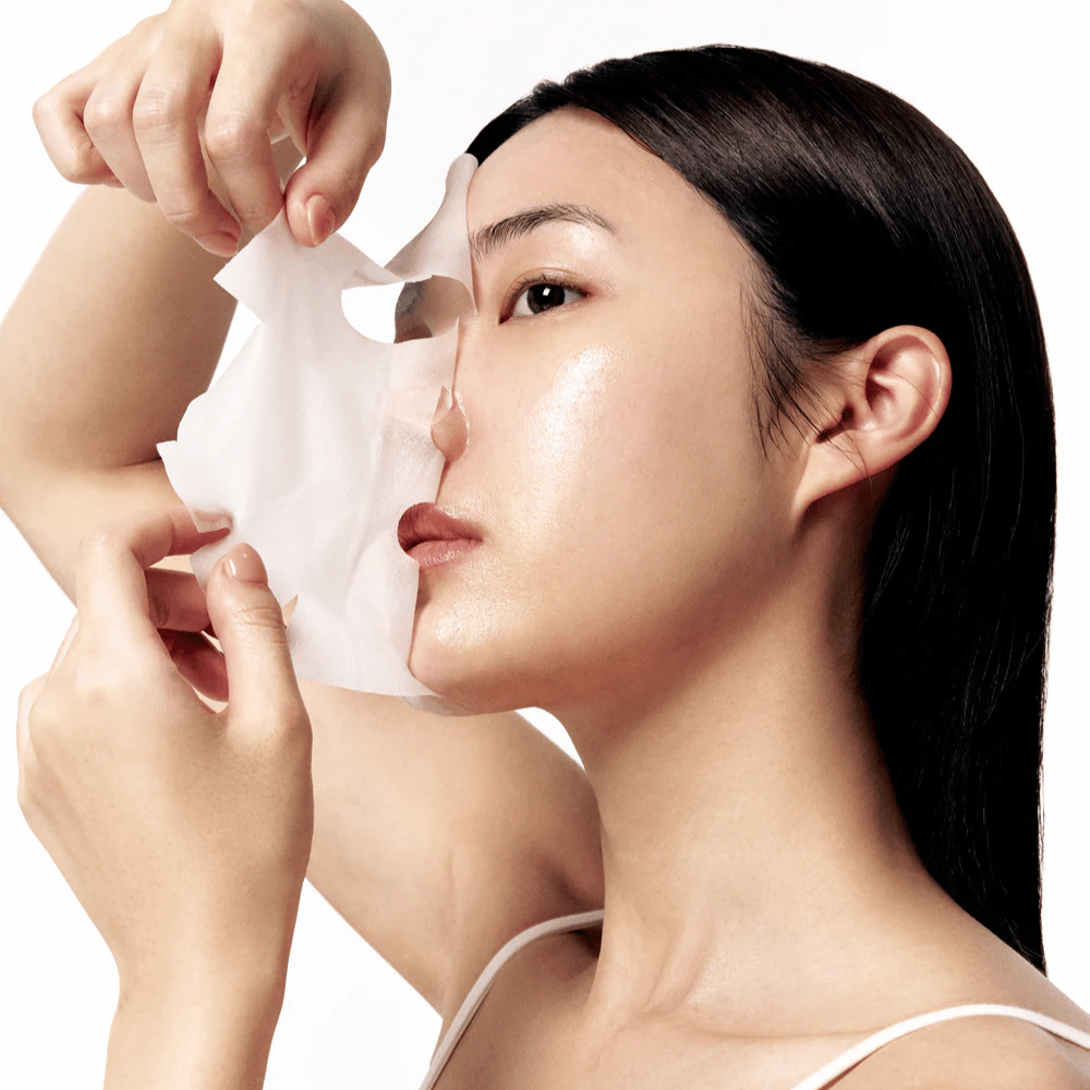 Kvinde med glat, fugtet hud fjerner en hvid Sheet mask fra ansigtet, hvilket afslører en strålende og jævn hudtone.