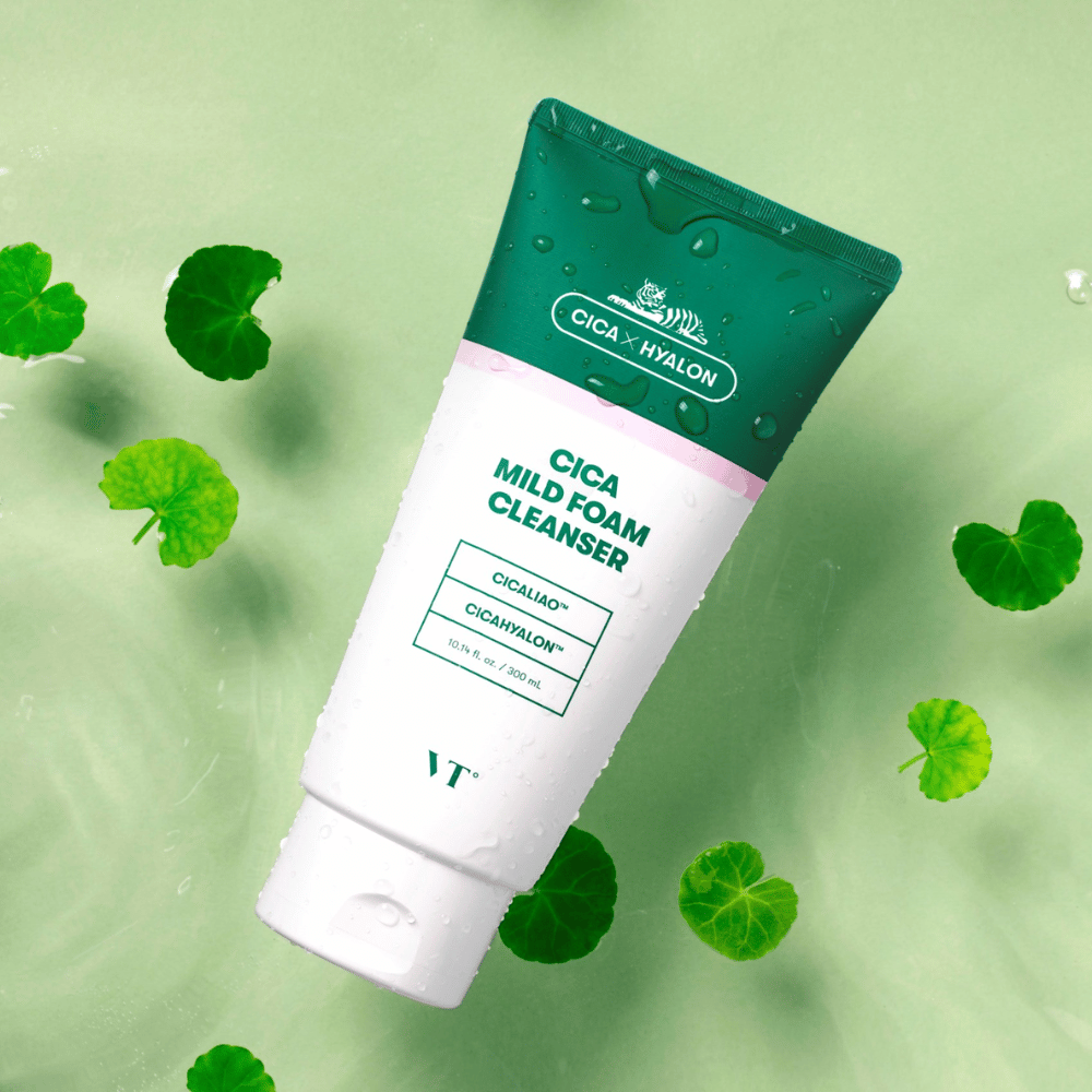 VT Cica Mild Foam Cleanser liggende mod en lysegrøn overflade med vanddråber på emballagen og omgivet af grønne centella asiatica-blade, hvilket fremhæver produktets beroligende egenskaber.