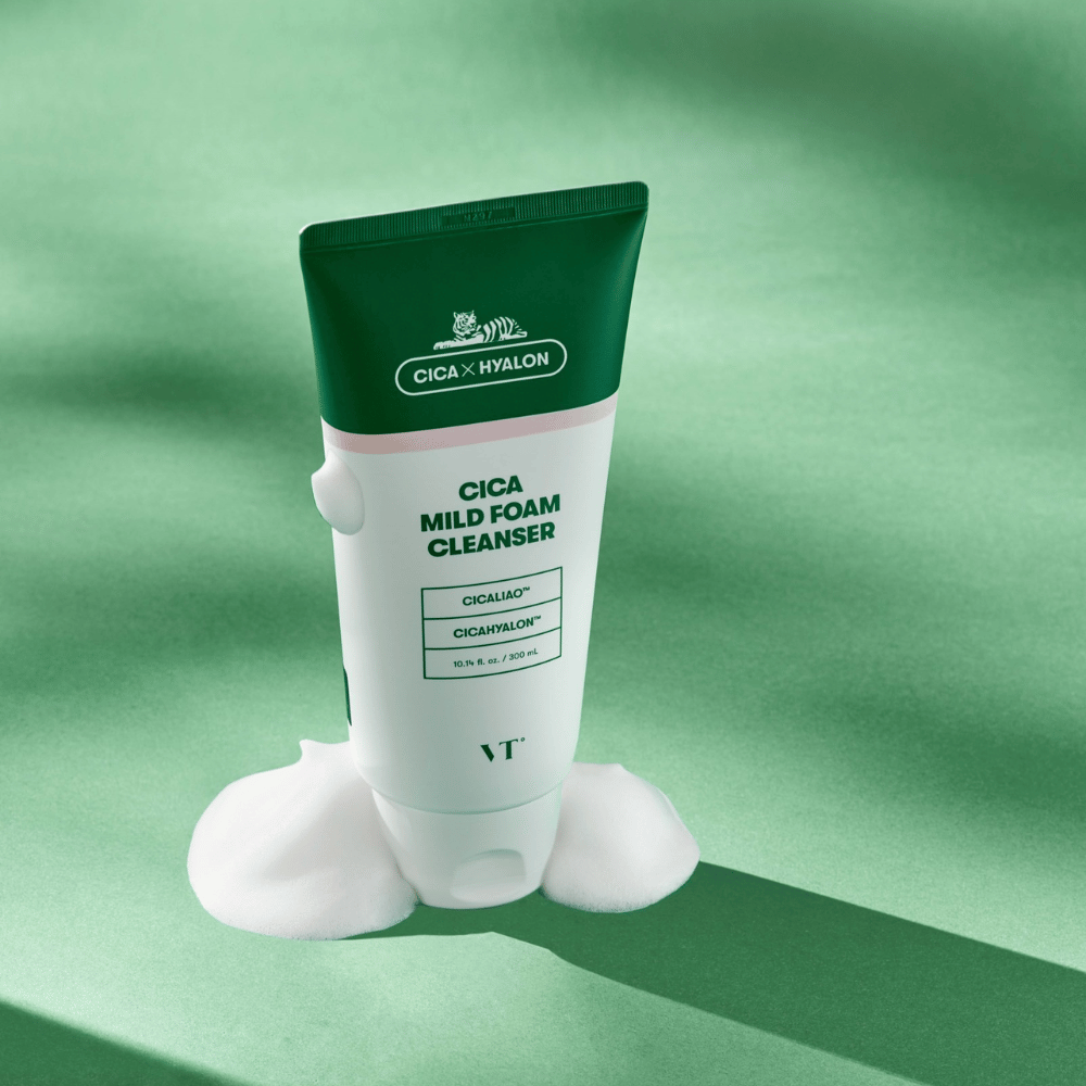 Tube med VT Cica Mild Foam Cleanser stående på en grøn overflade med skum rundt om bunden. Produktet har grøn og hvidt design og markedsføres med CICALIAO™ og CICAHYALON™.