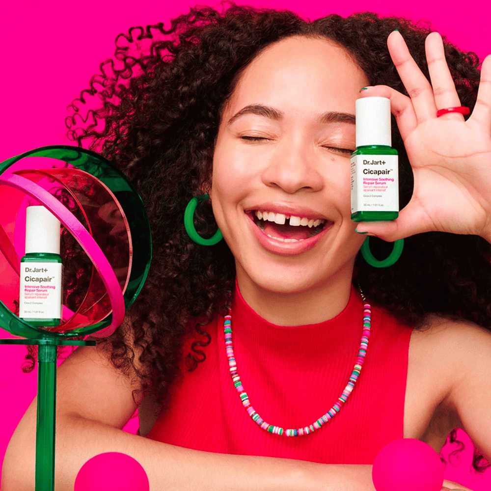 En kvinde smiler bredt, mens hun holder Dr.Jart+ Cicapair Intensive Soothing Repair Serum foran sit ansigt. Hun bærer grønne øreringe og en rød halskæde. Baggrunden er lyserød og dekoreret med grønne og lyserøde cirkler.