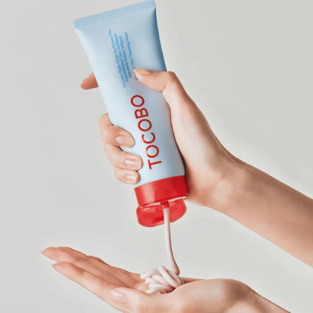 En person holder en tube af TOCOBO Coconut Clay Cleansing Foam. Tuben er lyseblå med en rød hætte og teksten "TOCOBO" på forsiden. Produktet trykkes ud af tuben og formes som en shampoo i hånden. Baggrunden er neutral og giver fokus på produktet.