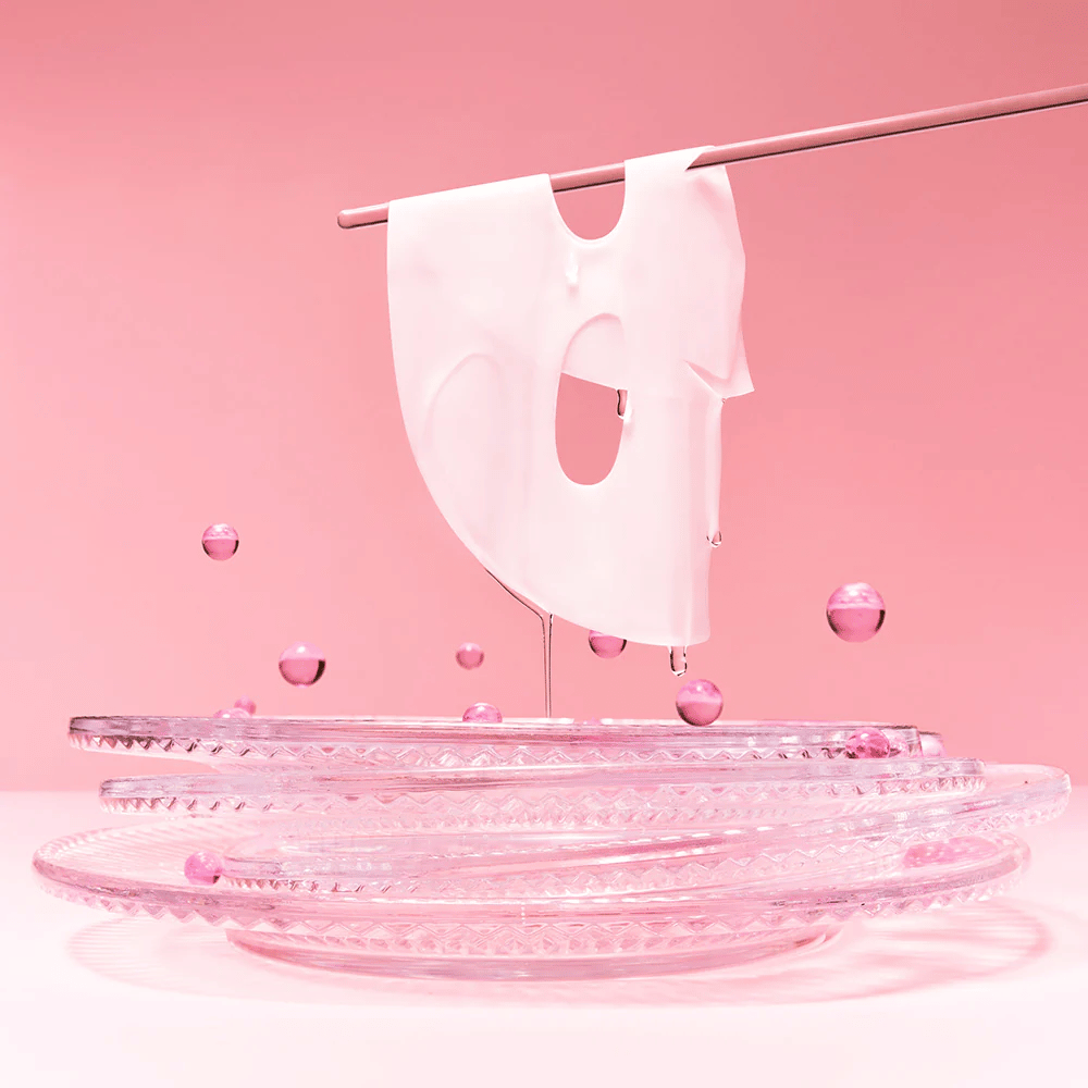 En hydrogelmaske hænger på en stang, dryppende med serum. Baggrunden er lyserød, og spejlet i bunden reflekterer de lyserøde bobler, der omgiver masken.
