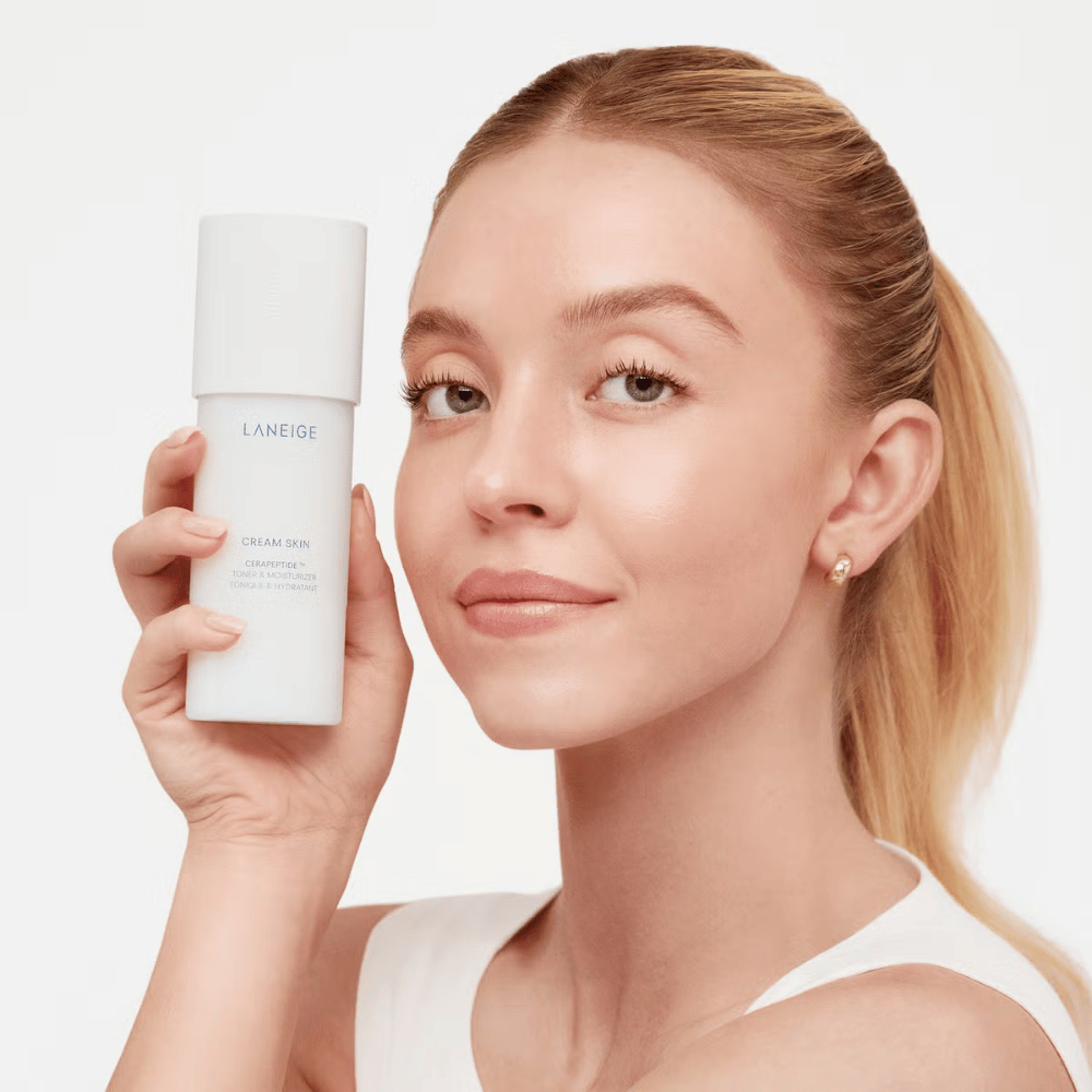 En person holder op Laneige Cream Skin Cerapeptide Refiner i en hvid flaske. Personen smiler og viser produktet, som markedsføres som en fugtgivende toner og hudplejeprodukt.