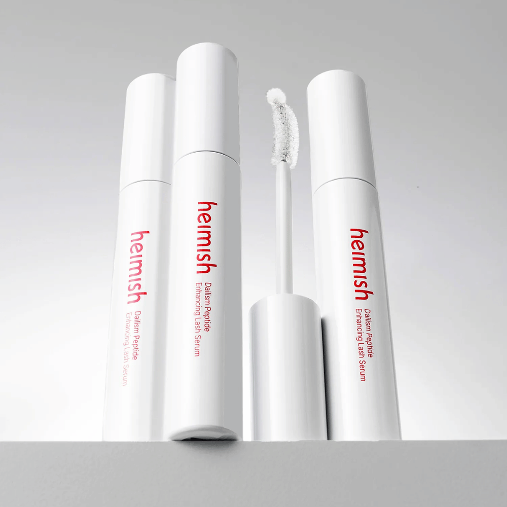 Flere pakninger af Heimish Dailism Peptide Enhancing Lash Serum vises sammen med den buede applikatorbørste. Billedet fremhæver produktets elegante design og præcisionsbørste til nem påføring.