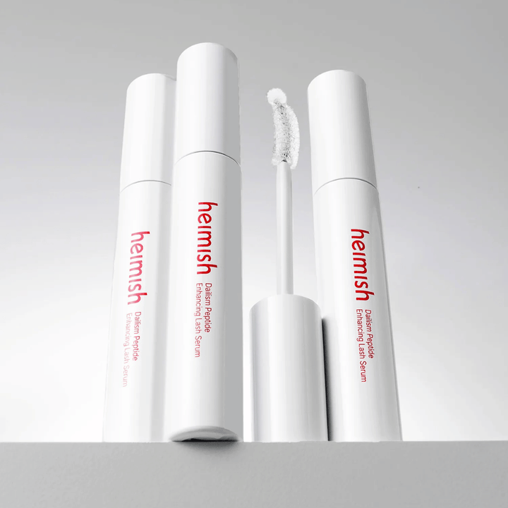 Flere pakninger af Heimish Dailism Peptide Enhancing Lash Serum vises sammen med den buede applikatorbørste. Billedet fremhæver produktets elegante design og præcisionsbørste til nem påføring.