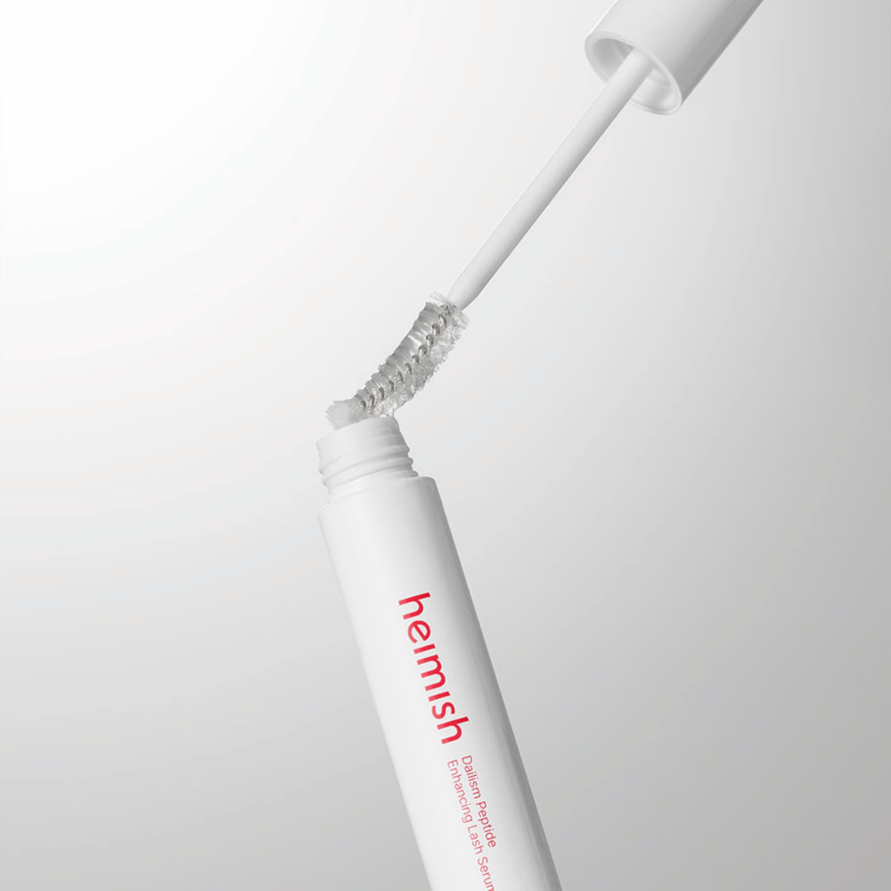 Nærbillede af applikatoren til Heimish Dailism Peptide Enhancing Lash Serum. Børsten trækkes op af den hvide beholder og viser den transparente serumformel, som påføres vipperne.