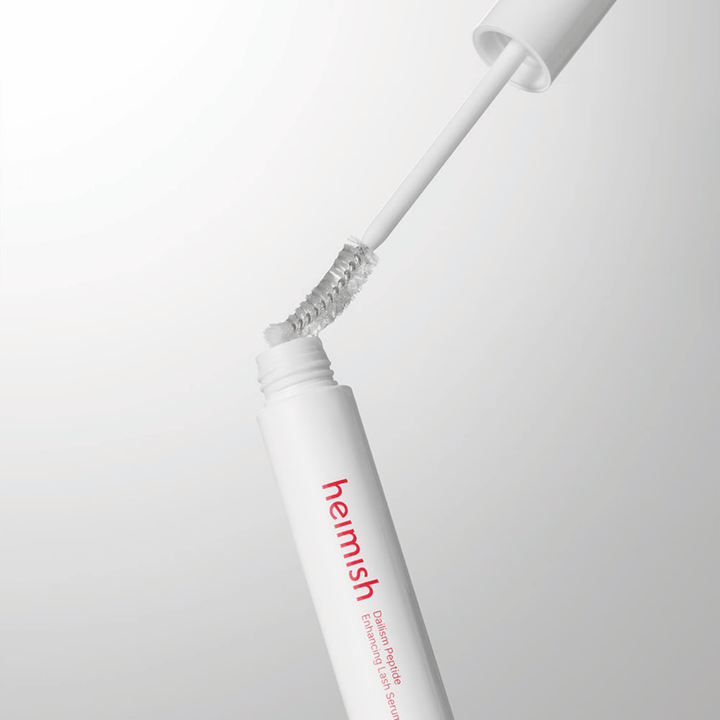 Nærbillede af applikatoren til Heimish Dailism Peptide Enhancing Lash Serum. Børsten trækkes op af den hvide beholder og viser den transparente serumformel, som påføres vipperne.