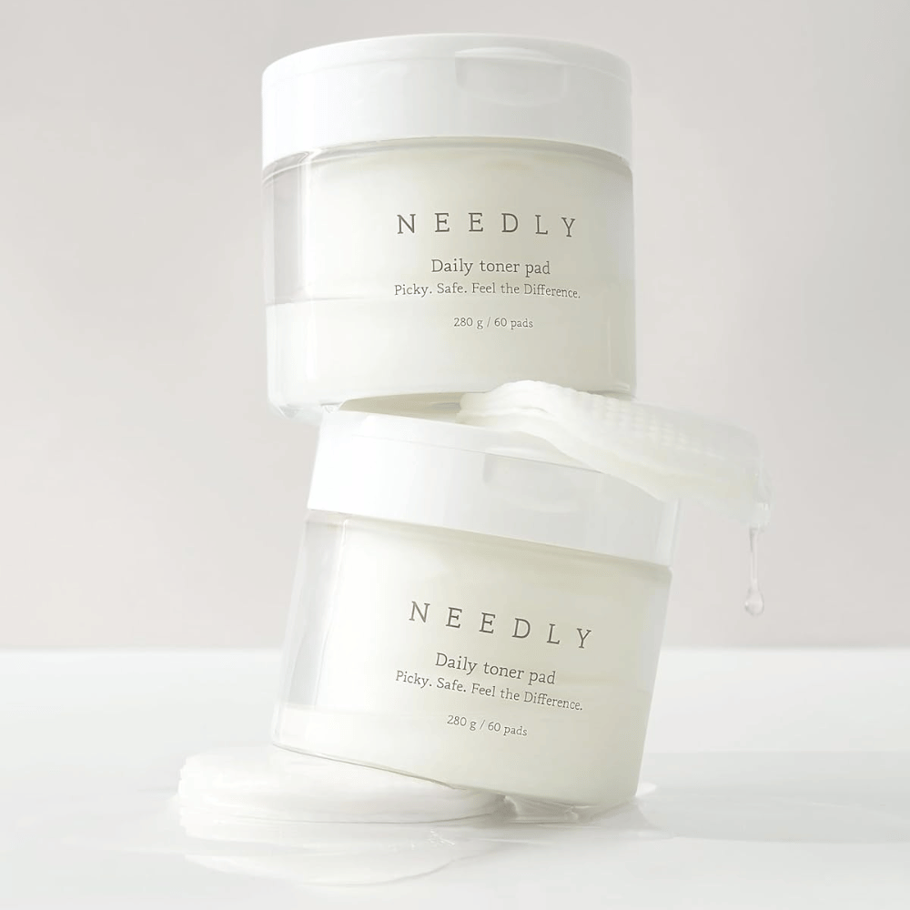 To krukker med Needly Daily Toner Pad stablet oven på hinanden. Krukkerne er hvide med minimalistisk tekst og indeholder 60 toner pads hver. En toner pad ligger delvist uden for den øverste krukke med væske, der drypper af.