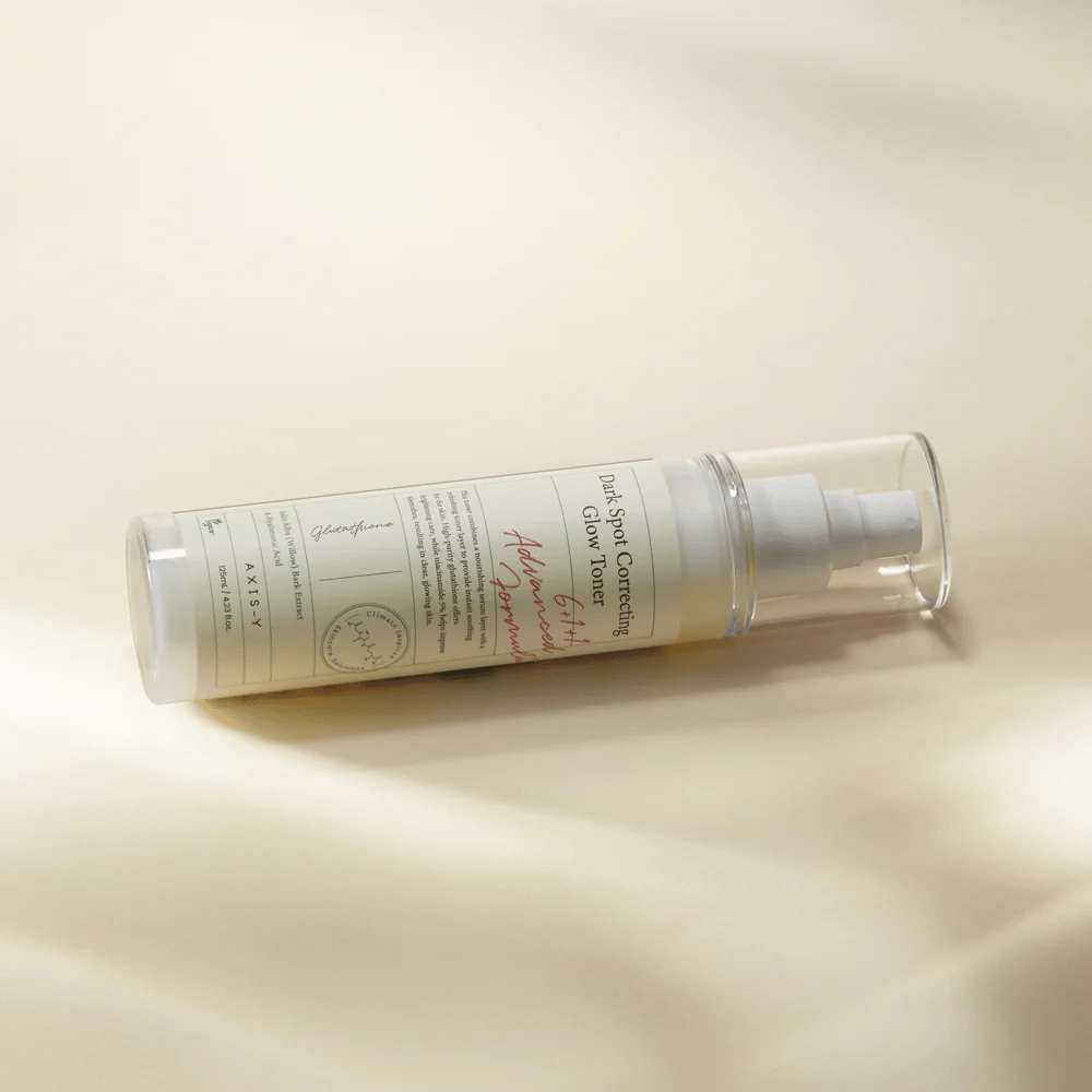 En liggende flaske af "AXIS-Y Dark Spot Correcting Glow Toner" placeret på en lys overflade. Flasken er gennemsigtig med en hvid etiket og har en hvid spraypumpe. Etiketten viser information om produktets lysnende og fugtgivende egenskaber samt indeholder nøgleingredienser som Glutathione og Niacinamide.