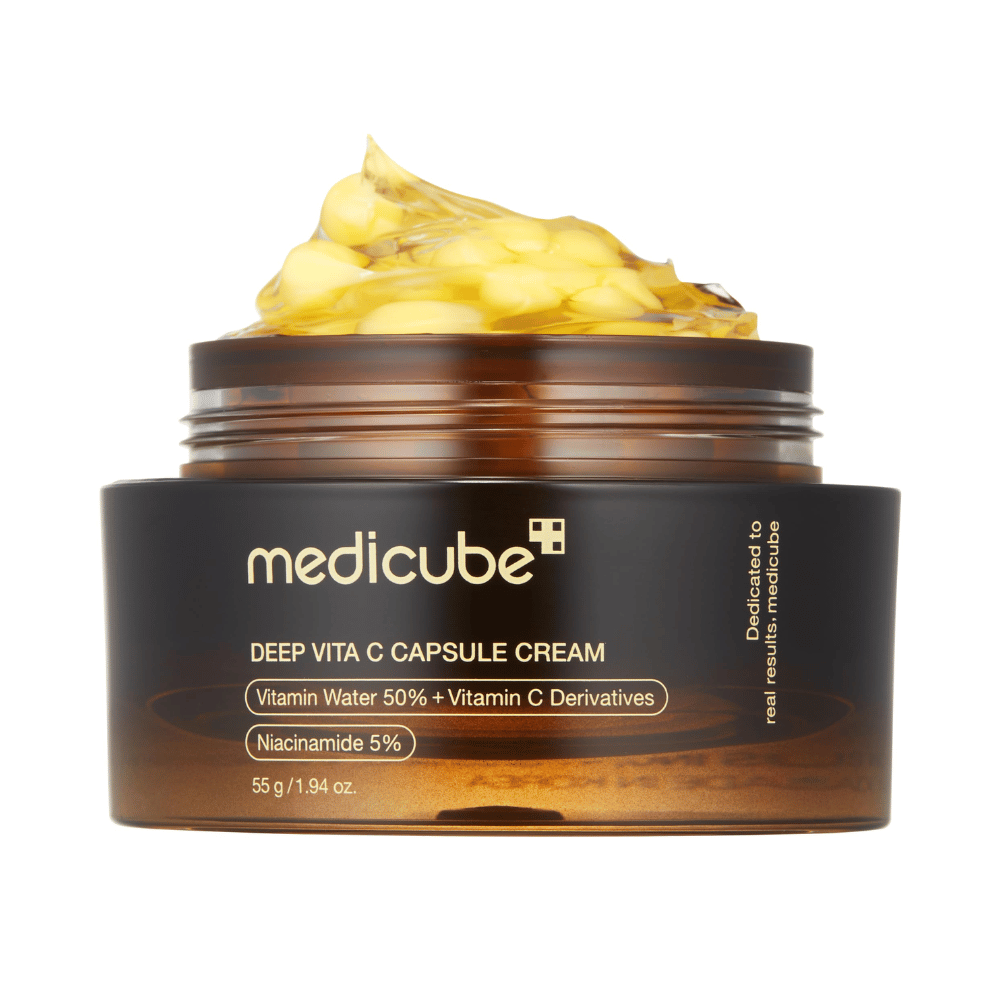 Et nærbillede af "Medicube Deep Vita C Capsule Cream," som viser kremens konsistens og kapslerne af vitamin C. Emballagen er mørkebrun med guldbogstaver og beskriver indholdet af vitaminvand (50 %) og niacinamid (5 %), hvilket giver en visuel præsentation af produktets fugtgivende og hudforbedrende egenskaber.
