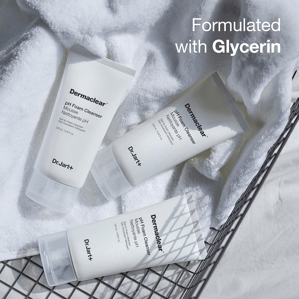 Tre tuber av Dr.Jart+ Dermaclear pH Foam Cleanser ligger på en vit frottéhandduk i en metallkorg, med texten "Formulated with Glycerin" i övre hörnet.
