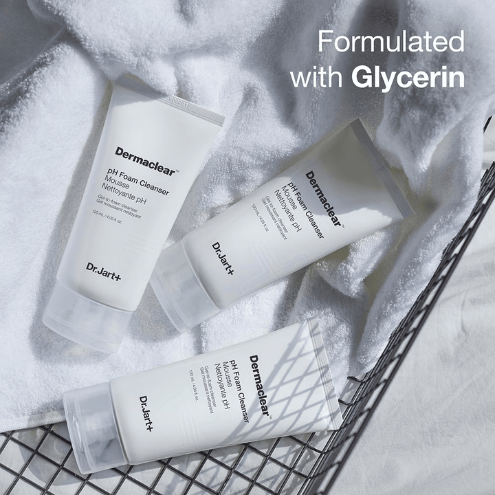 Tre tuber av Dr.Jart+ Dermaclear pH Foam Cleanser ligger på en vit frottéhandduk i en metallkorg, med texten "Formulated with Glycerin" i övre hörnet.