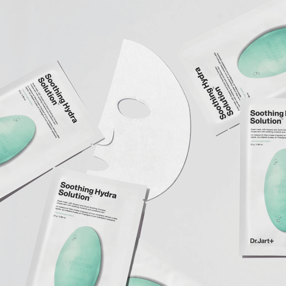 Flere pakker af "Soothing Hydra Solution" ansigtsmasker fra Dr.Jart+ er spredt ud på en lys overflade. En udfoldet maske er placeret i midten, hvilket viser dens hvide og hullede struktur.