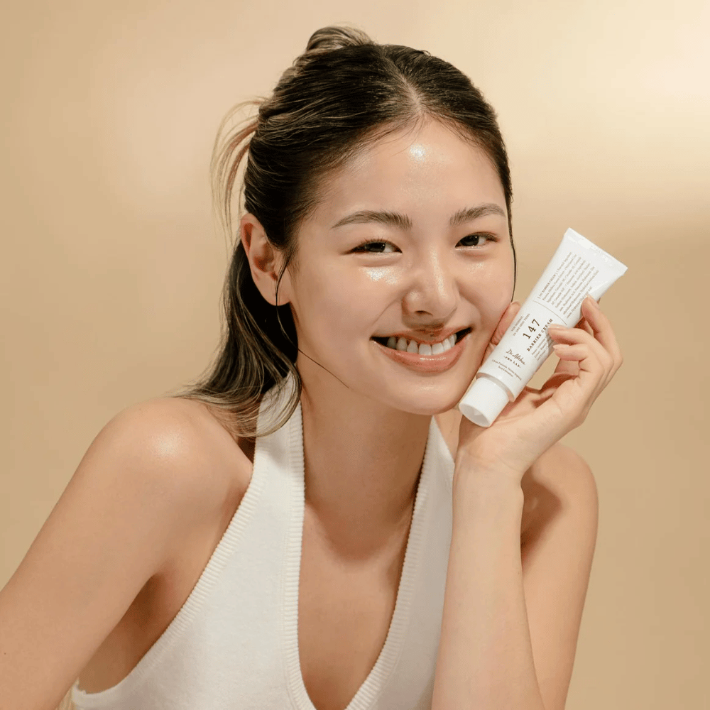 Model med glødende hud, som smiler og holder i Dr. Althea 147 Barrier Cream – en koreansk ansigtscreme til hudbarriere og fugtgivning.