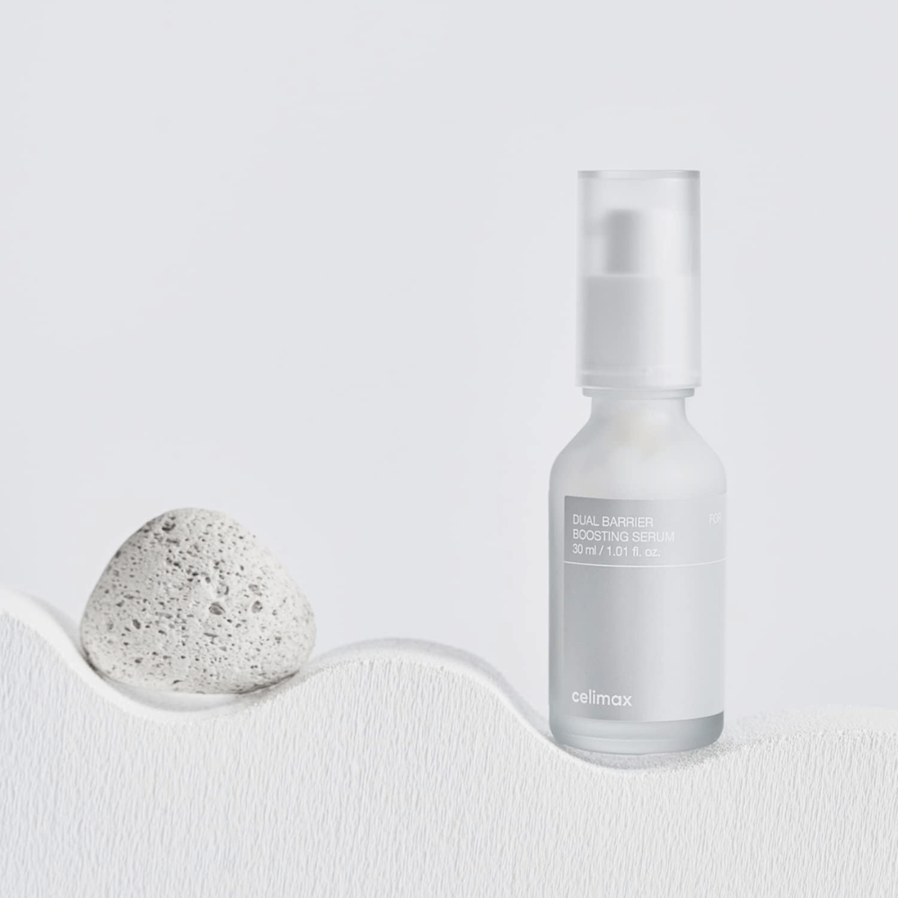 Celimax Dual Barrier Boosting Serum placeret på tekstil med sten, roligt produktbillede der signalerer fugt og styrket hudbarriere.
