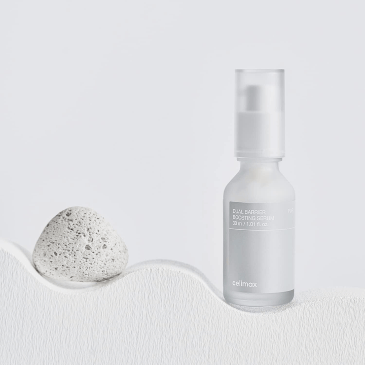 Celimax Dual Barrier Boosting Serum placeret på tekstil med sten, roligt produktbillede der signalerer fugt og styrket hudbarriere.