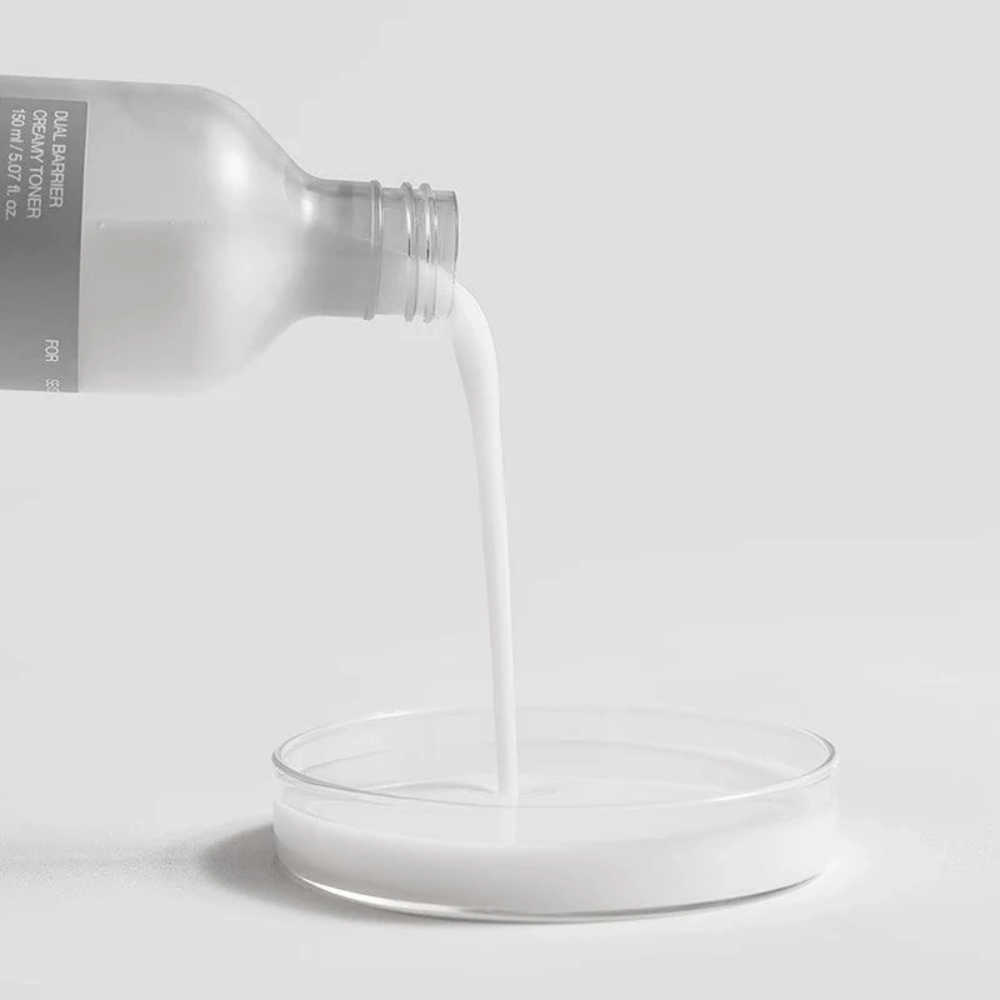 Celimax Dual Barrier Creamy Toner hældes forsigtigt fra flasken ned i en rund laboratoriebrønd, med sin mælkeagtige og næringsrige formel tydeligt i fokus.