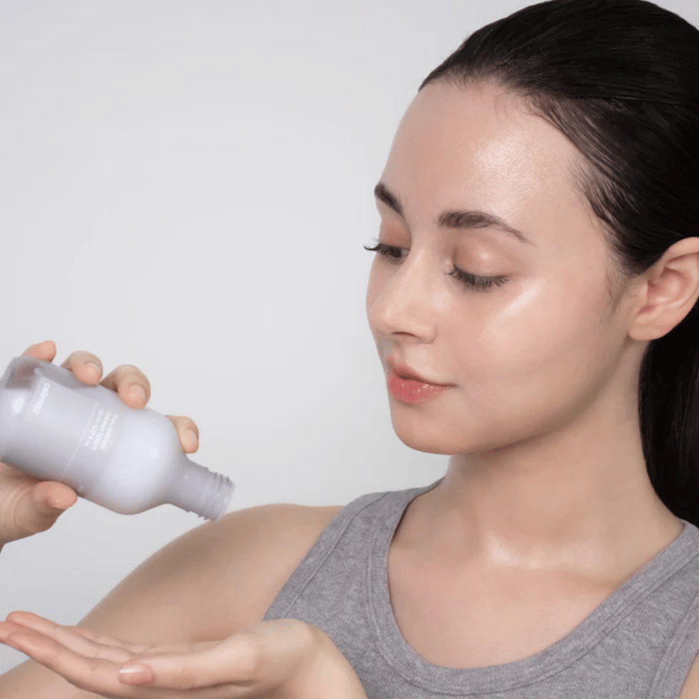 Kvinde med glansfuld, fugtet hud påfører Celimax Dual Barrier Creamy Toner ved at hælde produktet i håndfladen før sin hudplejerutine.