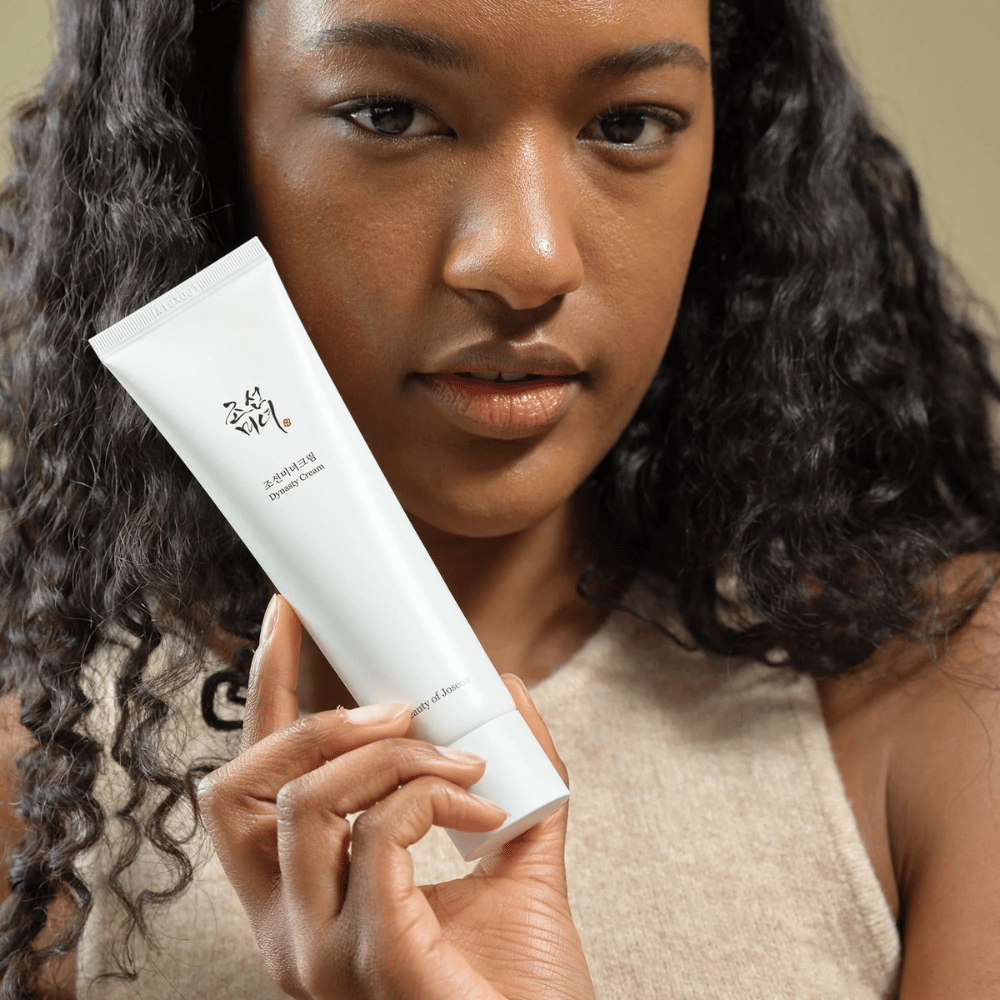 En kvinde med krøllet hår holder en tube "Beauty of Joseon Dynasty Cream" tæt på sit ansigt, hvilket fremhæver produktet. Cremen, præsenteret i en stilfuld hvid tube, er designet til at nære og forynge huden, hvilket understreger dens attraktivitet for forskellige hudtyper.