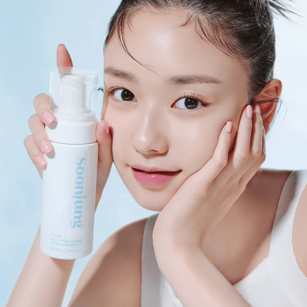 En ung kvinde med en klar hud holder en hvid pumpeflaske med teksten "Soon Jung pH 6.5 Whip Cleanser" fra ETUDE. Hun smiler til kameraet med en let makeup og en frisk udstråling mod en lyseblå baggrund.