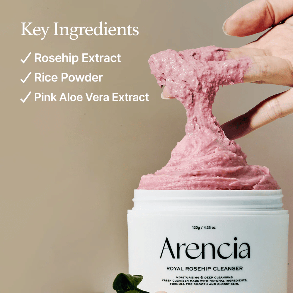 En hånd løfter den lyserøde, fluffy rensetekstur fra krukken Arencia Royal Rosehip Cleanser, med ingrediensliste til venstre: hybenekstrakt, rispulver og lyserød aloe vera-ekstrakt.