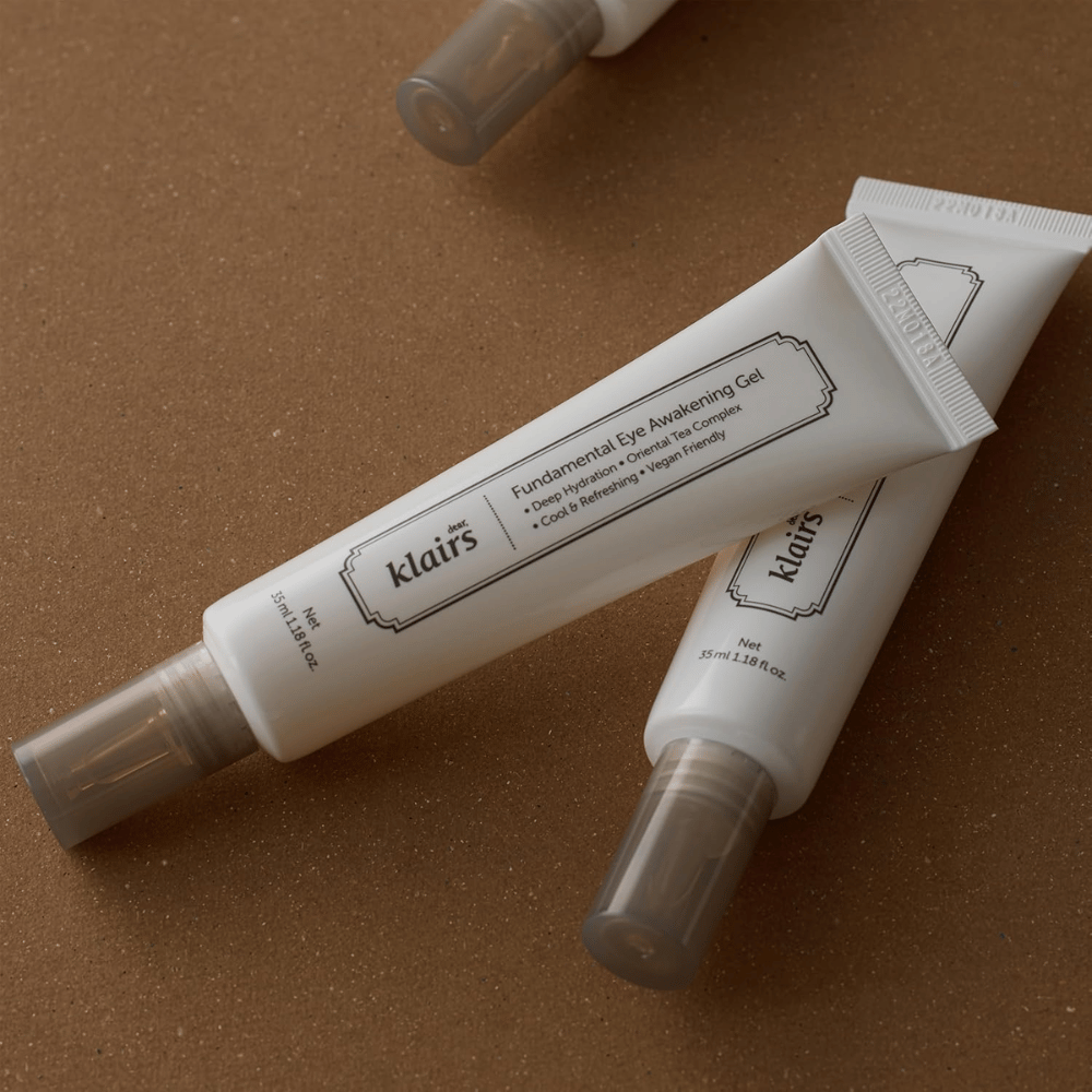 Lifestylebillede med to tuber af Klairs Fundamental Eye Awakening Gel placeret mod en varm brun baggrund. Kompositionen fremhæver produktets elegante design og premiumfølelse, samtidig med at fokus er på øjenpleje og fugt.