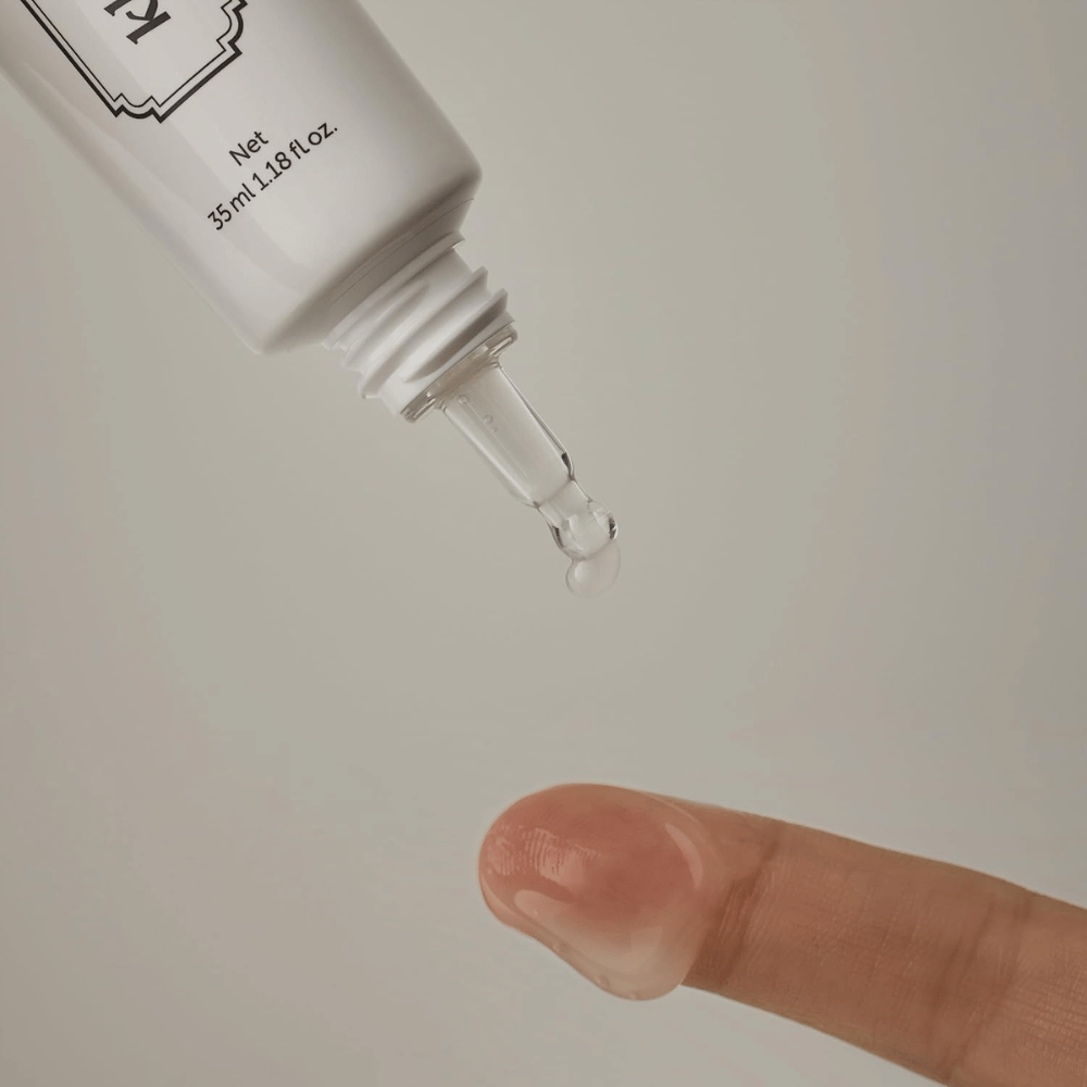 Detaljebillede, hvor en klar, let gel påføres fra den smalle spids mod en finger. Billedet illustrerer produktets transparente, gelbaserede konsistens og den præcise påføring omkring øjenområdet.