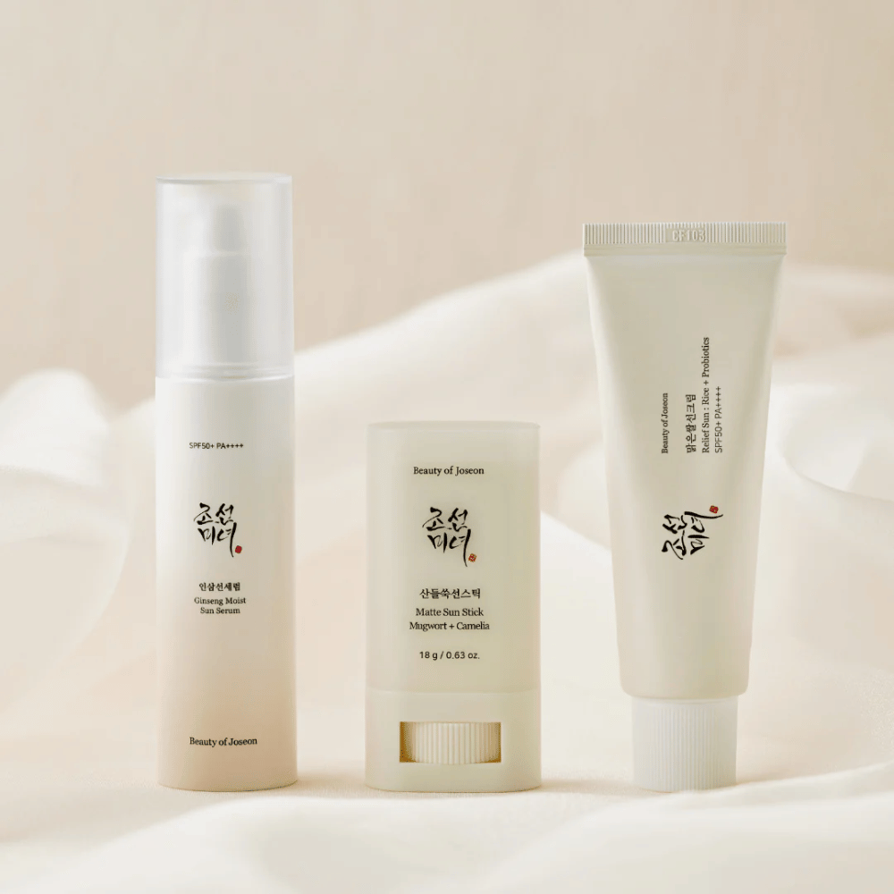 Tre solbeskyttelsesprodukter fra Beauty of Joseon – Ginseng Moist Sun Serum, Matte Sun Stick og Relief Sun Rice + Probiotics – står opstillet på et cremefarvet stof med blødt lys.