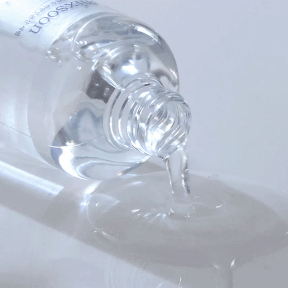 Nærbillede af gennemsigtigt serum, der hældes fra en flaske Mixsoon Glacier Water Hyaluronic Acid Serum på en blank overflade.