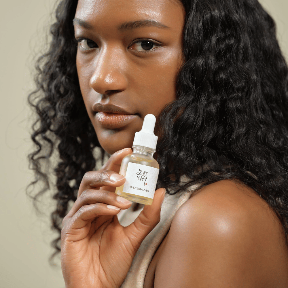 Kvinde med krøllet hår holder en flaske Beauty of Joseon Glow Serum med pipette, en fugtgivende og glødgivende hudplejeprodukt.