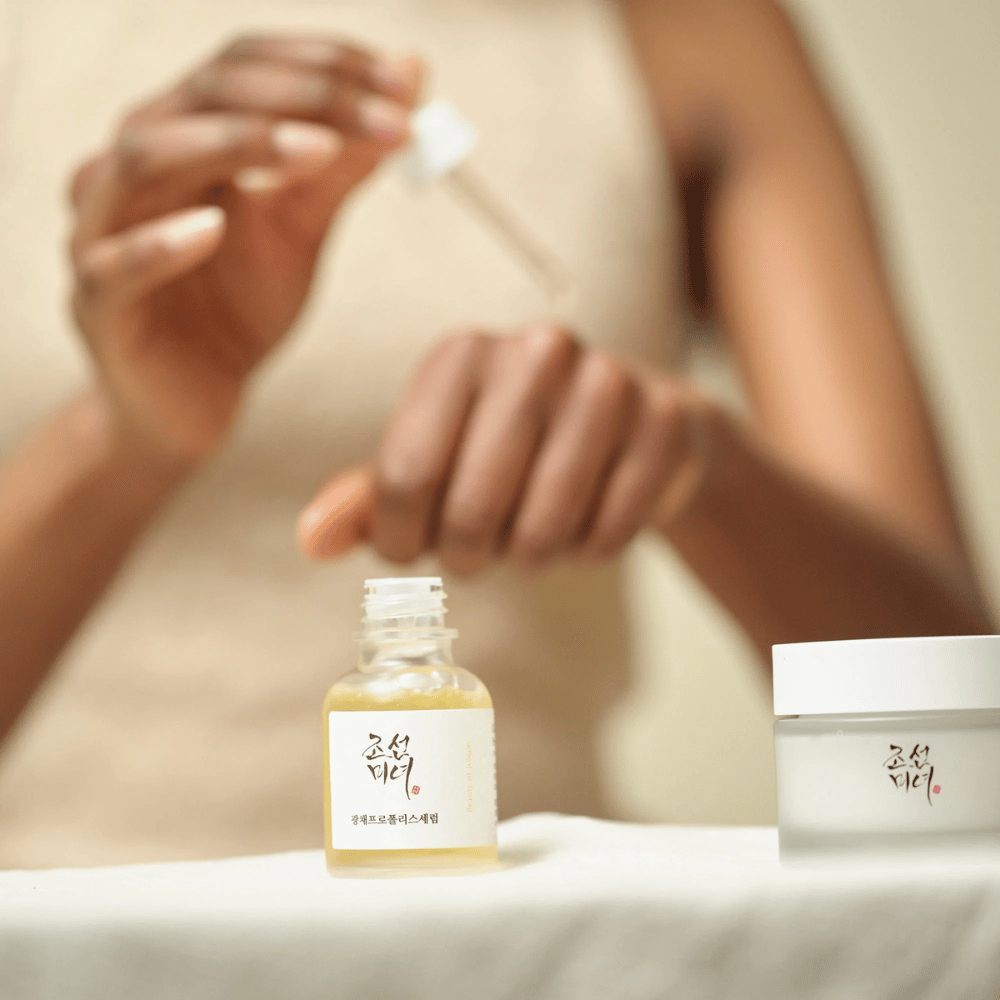 Person der påfører Beauty of Joseon Glow Serum med pipette fra glasflaske, med en krukke af samme serie ved siden af på bordet.