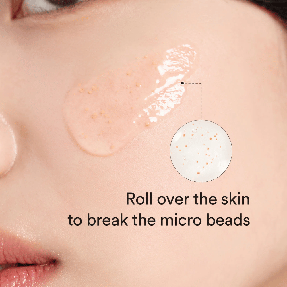 Nærbillede af en kind med et serum indeholdende små mikrokugler påført huden. En cirkelzoom viser kuglerne tydeligere. Teksten siger “Roll over the skin to break the micro beads”.