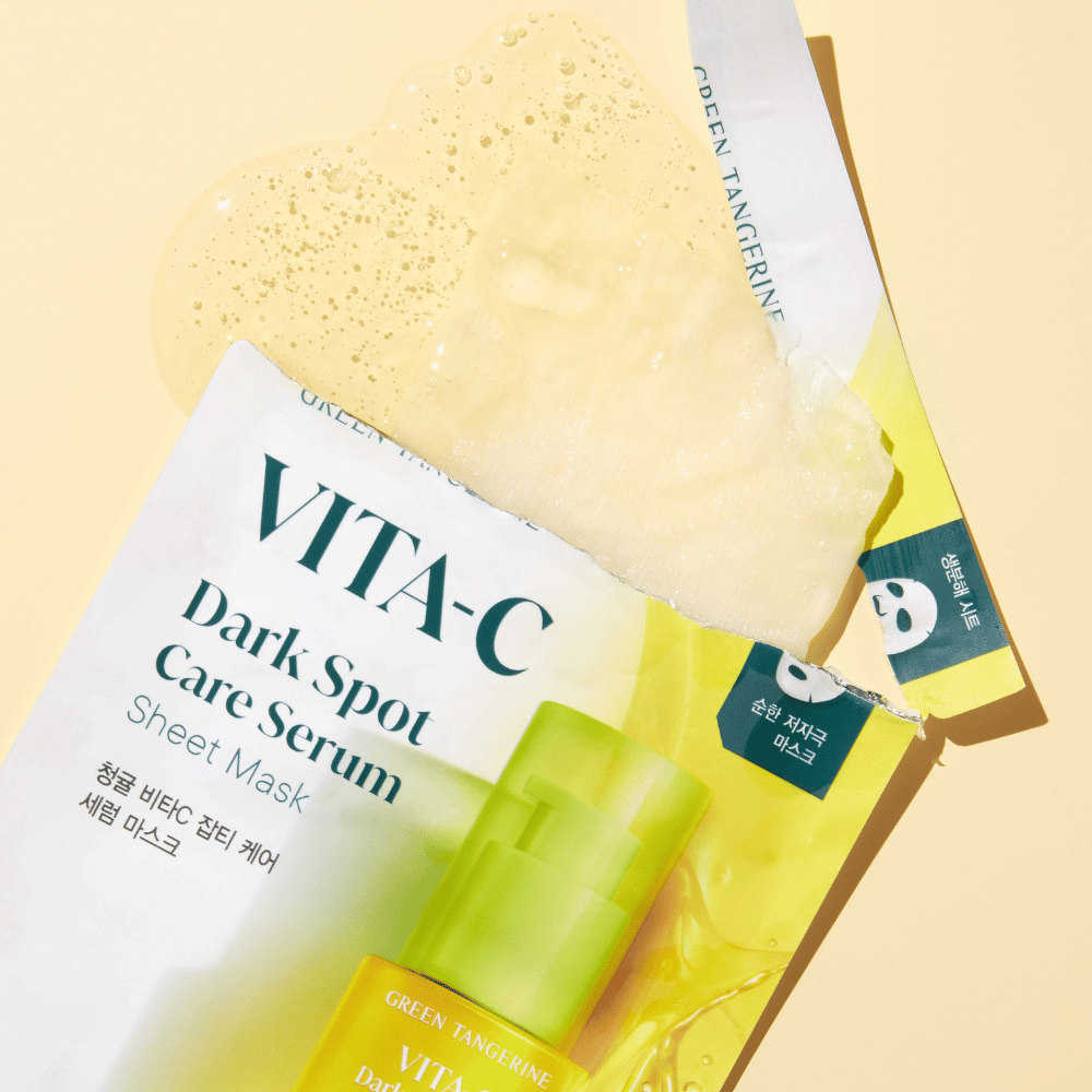 Åben emballage af VITA-C Dark Spot Care Serum Sheet mask med en ansigtsmaske ovenpå og en grøn serumflaske, alle mod en gul baggrund.