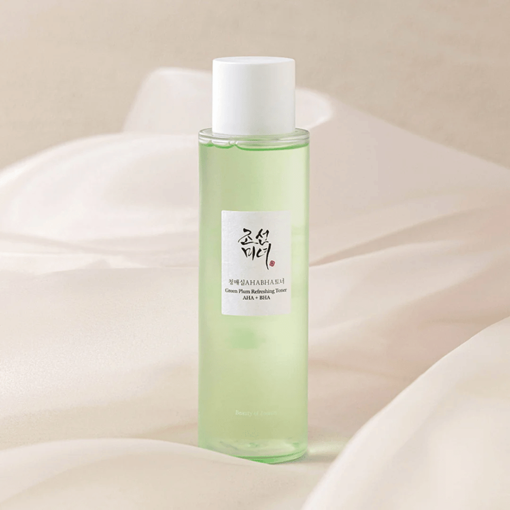 En flaske med "Green Plum Refreshing Toner" AHA + BHA står på en blød, lys baggrund. Produktet har en grøn væske, der afspejler friskhed og naturlighed. Etiketten er hvid med sorte og røde indslag, hvilket giver et elegant og rent look.