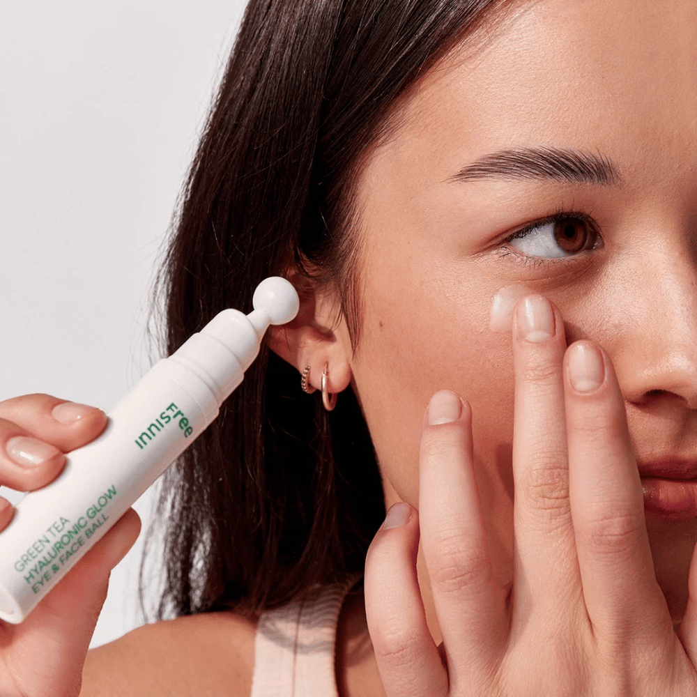 En person anvender Innisfree Green Tea Hyaluronic Glow Eye & Face Ball på sin hud under øjnene. Produktet har en hvid, rund applikationsdel og er i en hvid flaske med grøn tekst. Brugerens ansigt ses delvist og fokuserer på anvendelsen af produktet.