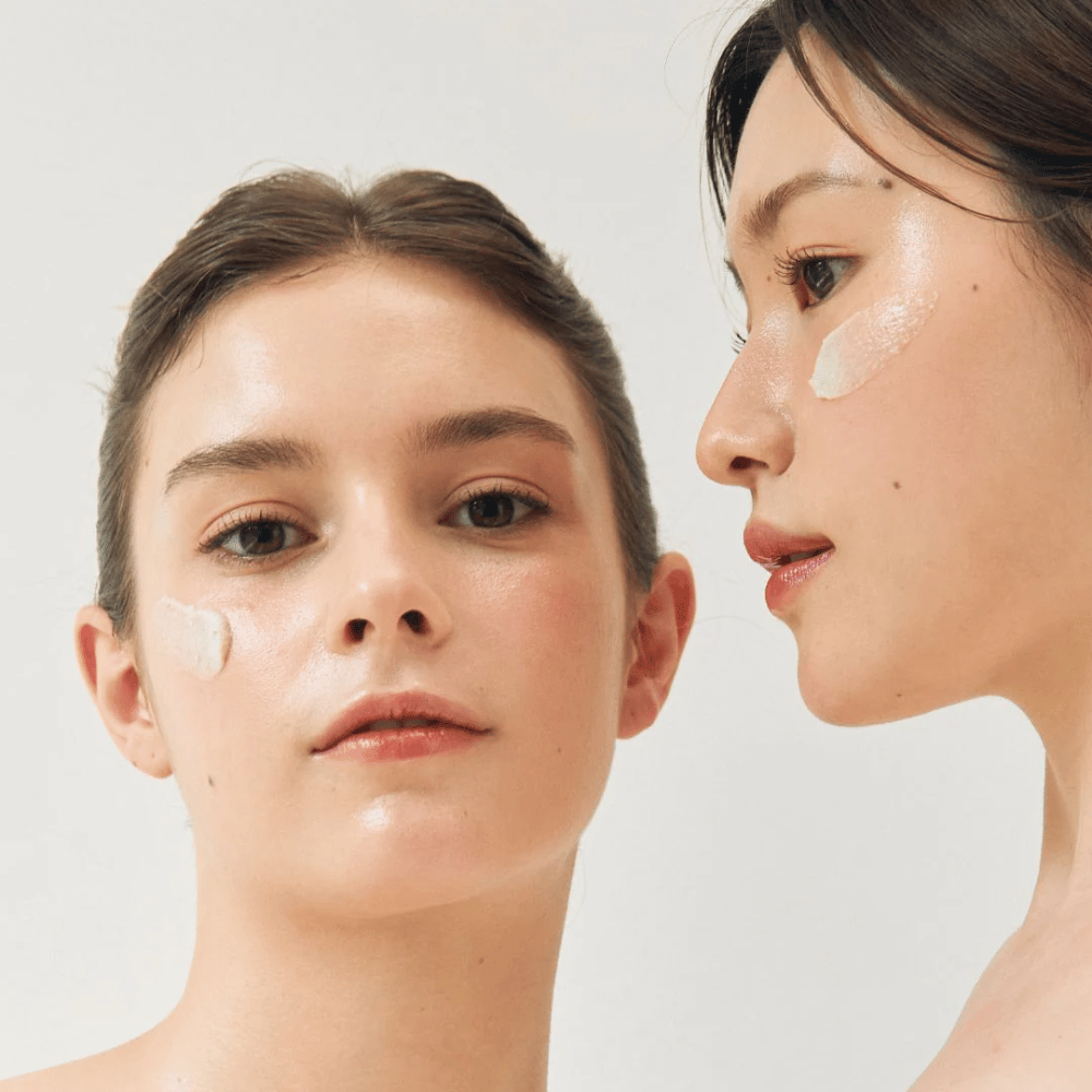 To kvinder med ansigtsmasker påført dele af deres ansigter. Den ene kvinde har maske på kinden og næsen, mens den anden har den under øjet og på kinden. De har naturlig makeup og ser friske ud, hvilket fremhæver maskernes fugtgivende og lysende effekt.