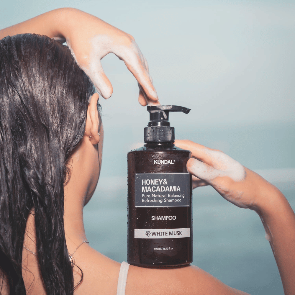 Kvinde med vådt hår påfører skum og holder KUNDAL Honey & Macadamia Nature Shampoo White Musk mod havbaggrund.