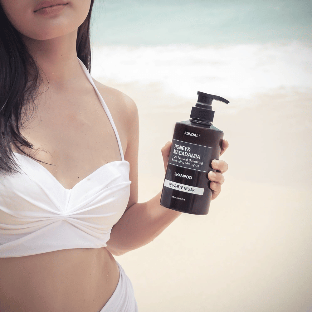 Kvinde i hvid bikini holder KUNDAL Honey & Macadamia Nature Shampoo White Musk på en strand med havet i baggrunden.