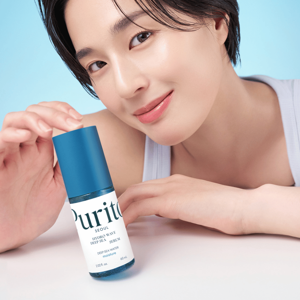 En kvinde med kort hår holder et blåt serum fra Purito, Hydro Wave Deep Sea Serum. Hendes hud ser strålende og fugtig ud, hvilket fremhæver produktet i fokus. Baggrunden er lyseblå, hvilket giver en frisk og ren følelse. Produktet er fordybet i hudpleje.