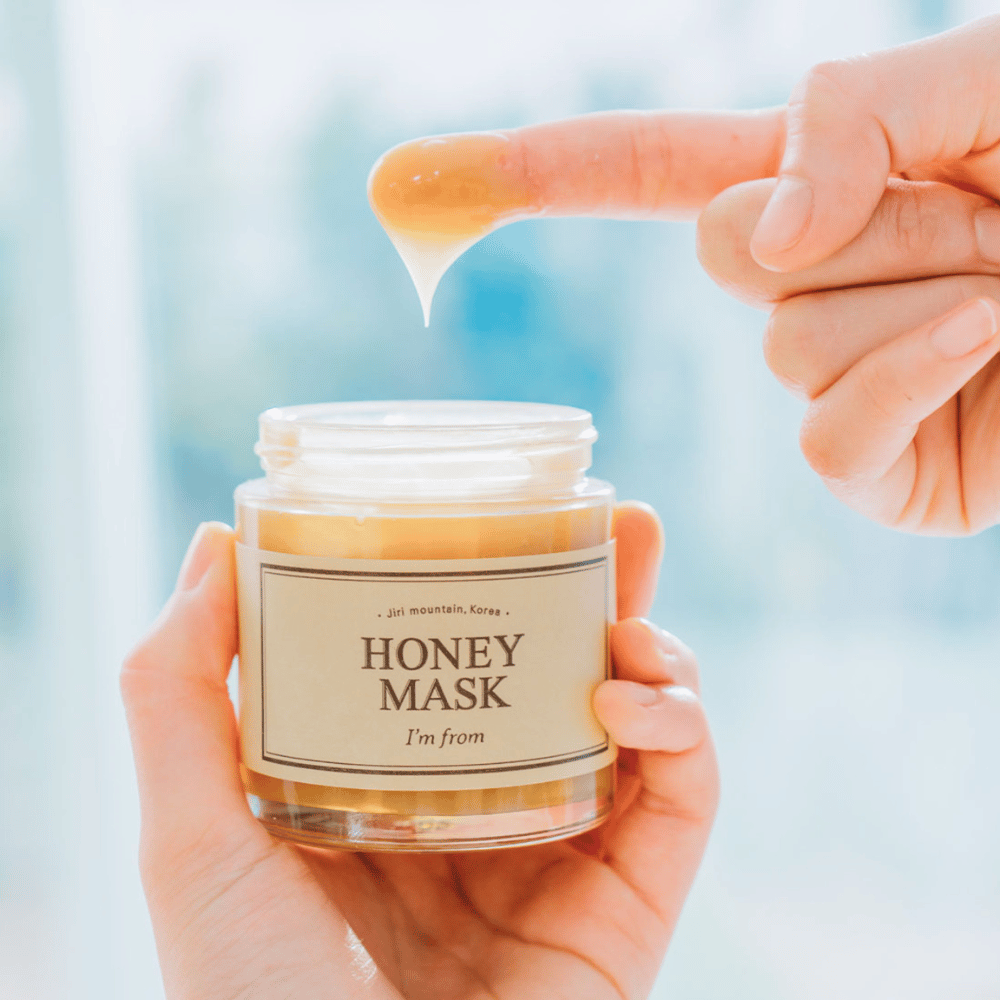 En hånd holder åben krukke af "I'm from Honey Mask", og en finger dyppes i den gyldne maske mod en lys baggrund.