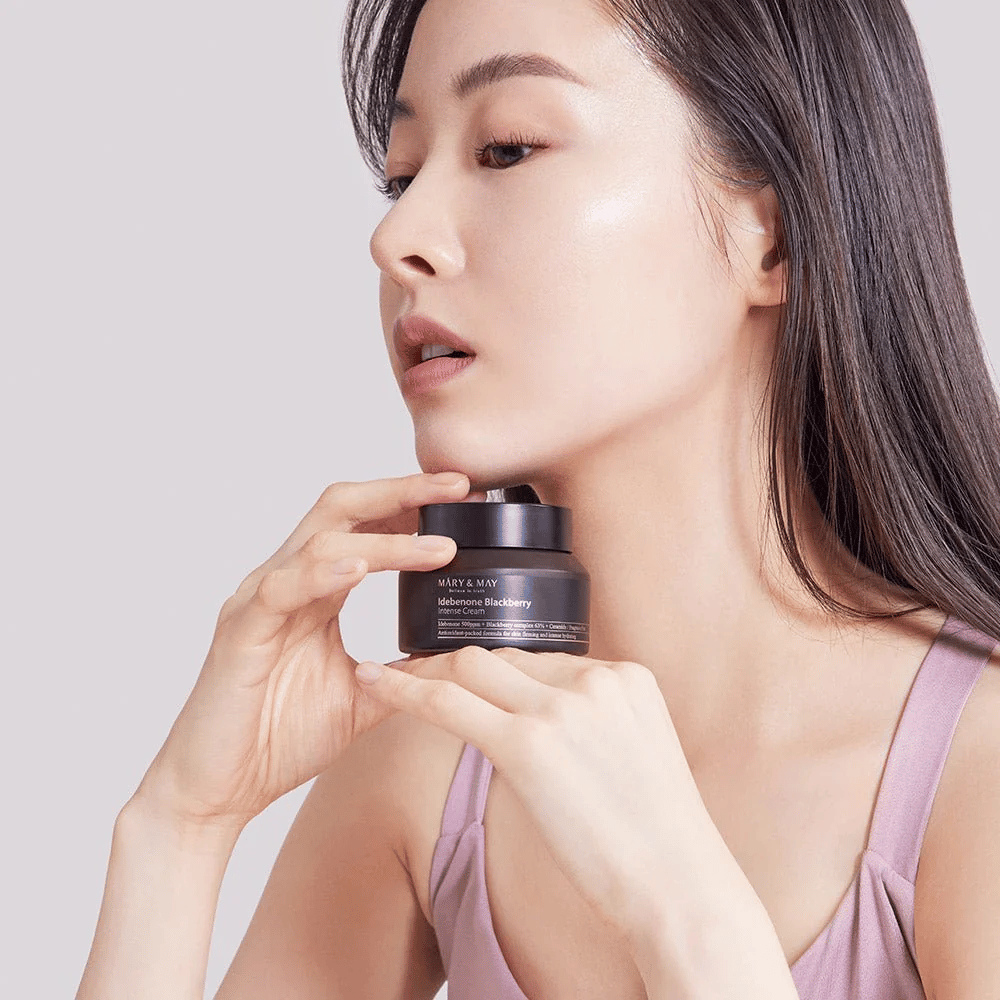 En kvinde holder en krukke med MARY & MAY Idebenone Blackberry Intense Cream. Hun har en strålende hud og smiler let, klædt i en ærmeløs top. Produktet er en intensiv creme med idebenon og brombærkompleks, som er beregnet til hudens fasthed og fugt.