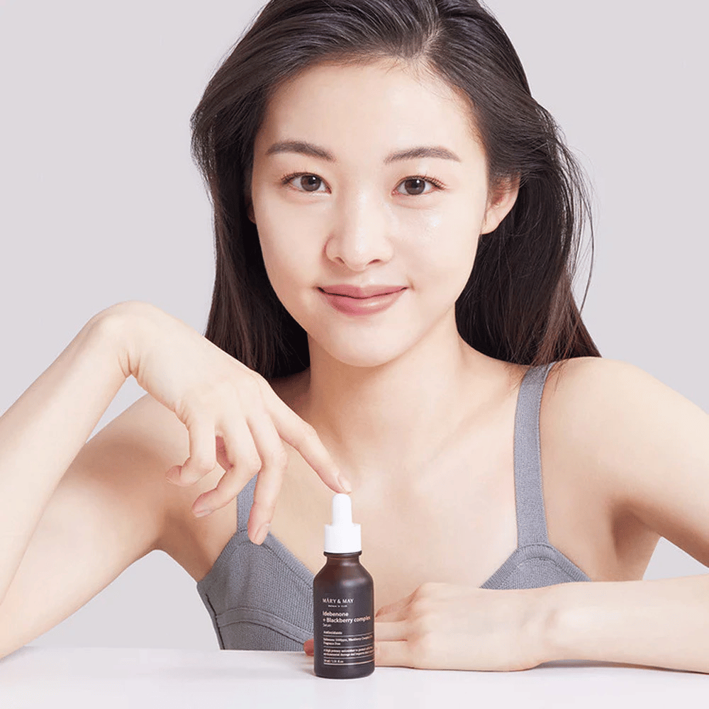 En kvinde med langt, mørkt hår holder i MARY & MAY Idebenone + Blackberry Complex Serum, en mørk glasflaske med pipette. Hun har et glad smil og peger på flasken med sin anden hånd. Kvinden er klædt i en grå, ærmeløs top og ser ud til at befinde sig i et lyst og enkelt miljø, hvilket fremhæver produktets fokus på hudpleje og skønhed.