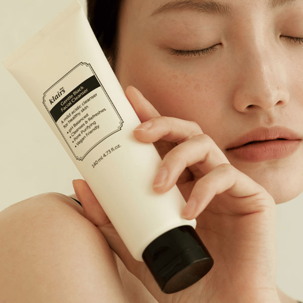 En person holder et rør af Klairs Gentle Black Facial Cleanser mod sin kind, lukkede øjne og naturlig hudtone ses i baggrunden.