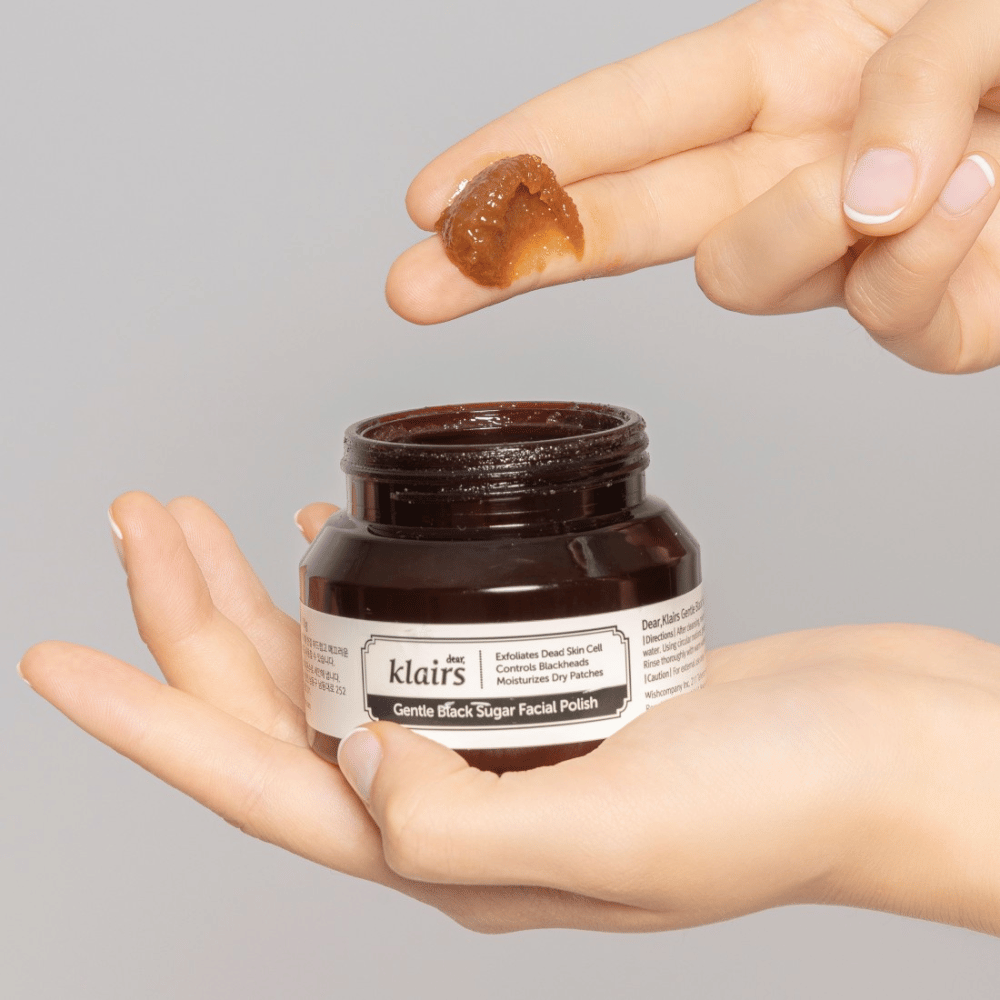 Hænder holder en åben krukke af Klairs Gentle Black Sugar Facial Polish og viser produktet på en finger, mod en grå baggrund.