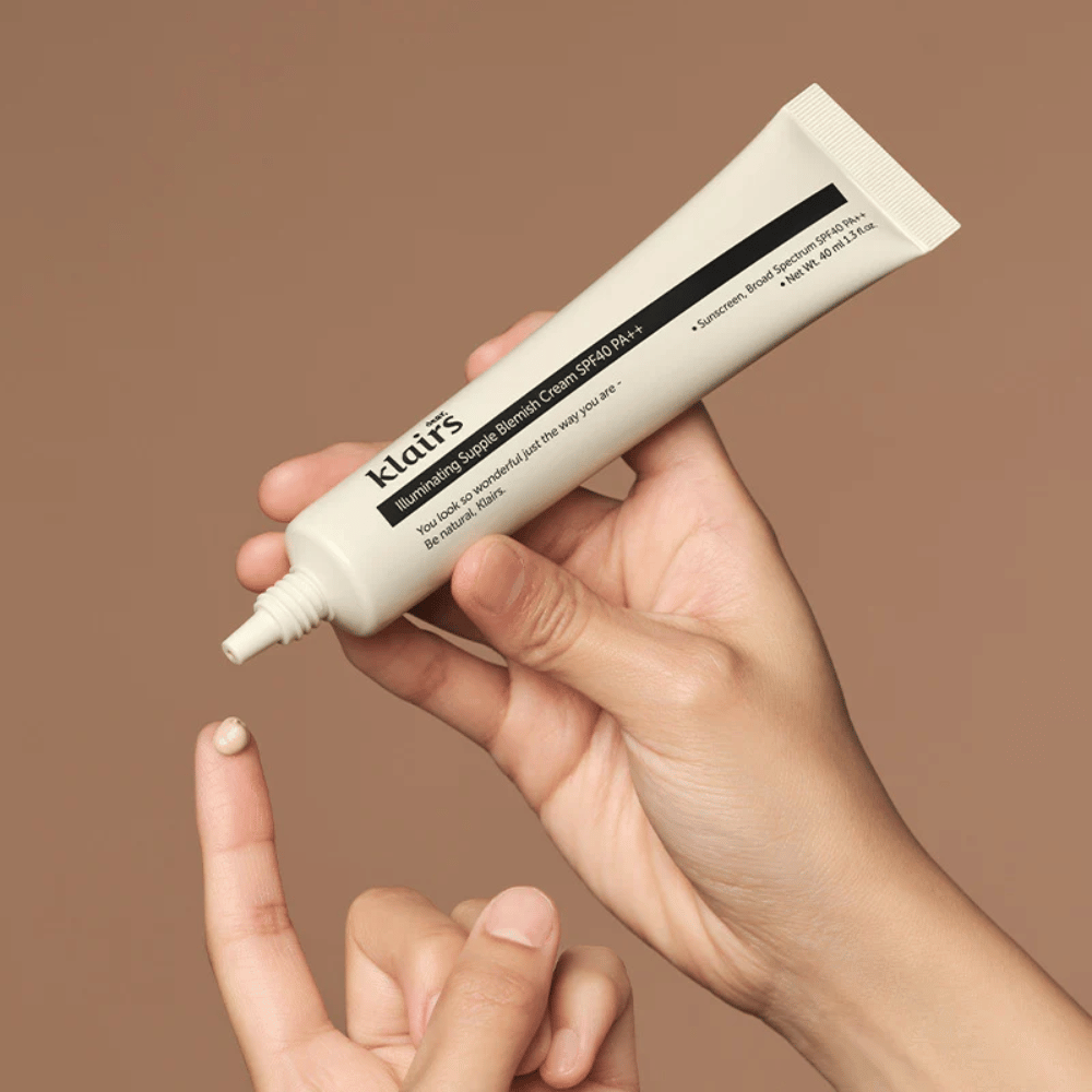 En hånd holder et rør af Klairs Illuminating Supple Blemish Cream SPF40 PA++ og trykker en lille mængde creme ud på en finger, mod en brun baggrund.