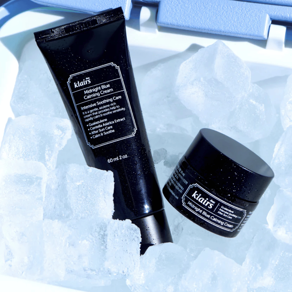 En tube og krukke af Klairs Midnight Blue Calming Cream ligger på isterninger, med fokus på beroligende og fugtgivende egenskaber, mod en blå baggrund.