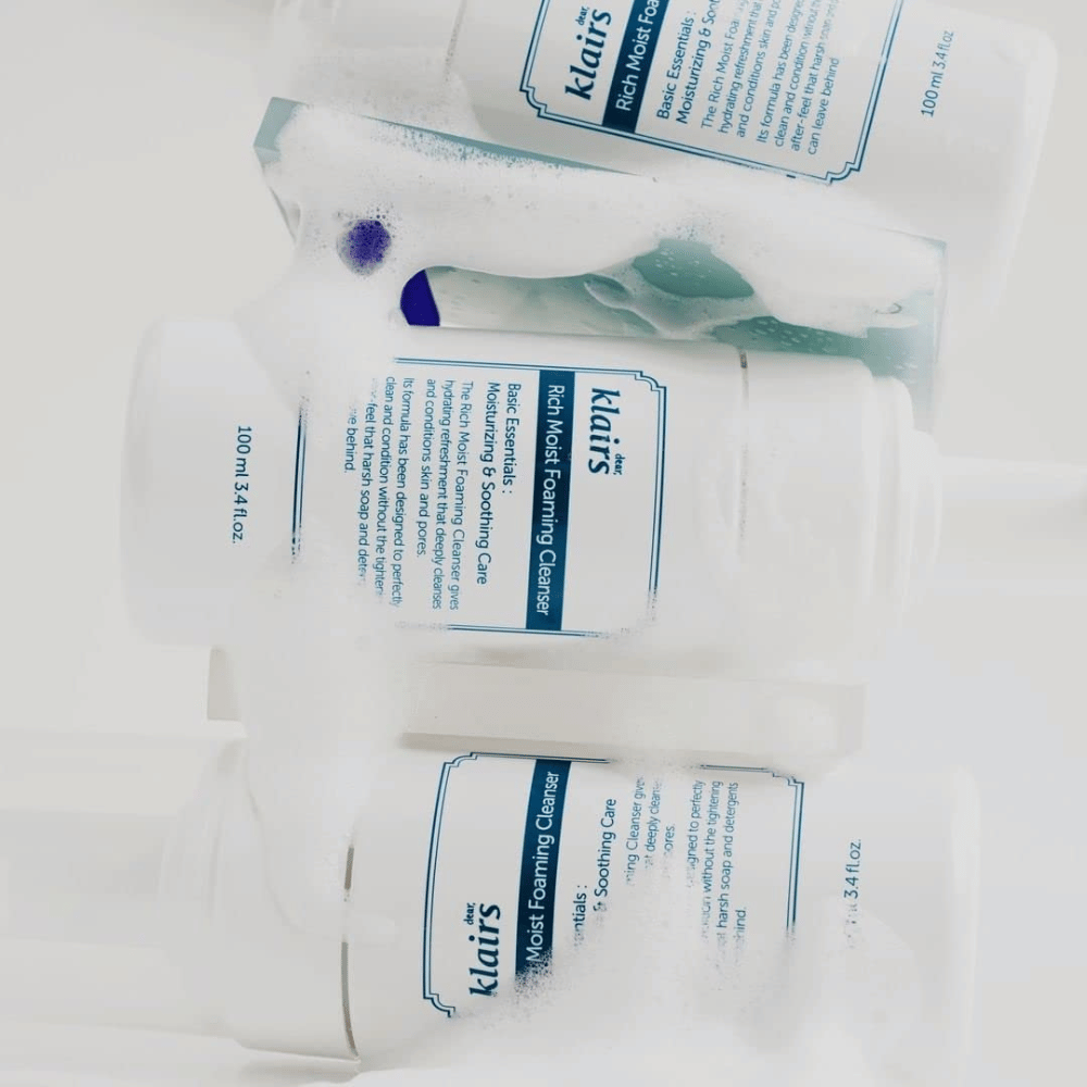 Tre flasker af Klairs Rich Moist Foaming Cleanser dækket med skum og vanddråber, placeret på en hvid overflade.