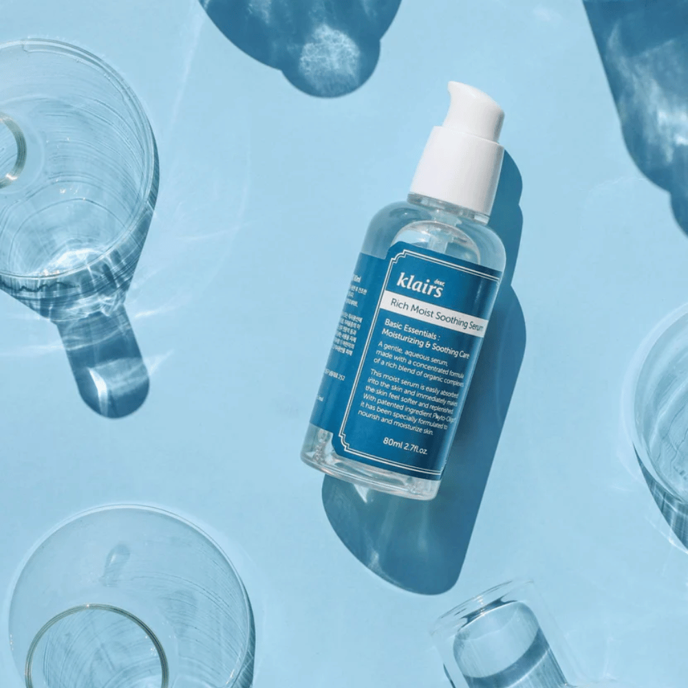 En flaske Klairs Rich Moist Soothing Serum blandt vandglas med refleksioner på en blå baggrund.